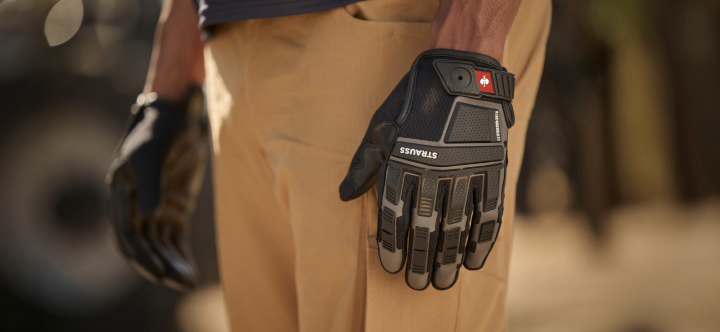 A close-up view shows a person wearing black protective work gloves with reinforced palms while standing next to tan work pants.