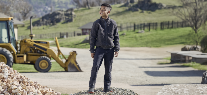 A young boy wearing dark work clothing stands on a gravel pile at a construction site with a yellow excavator in the background.