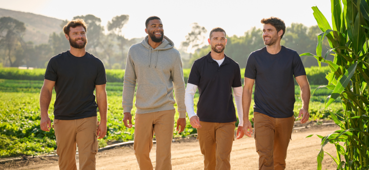 Four men wearing casual work clothing walk together through a green farm field on a sunny day.