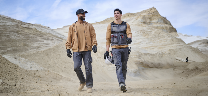 Two men wearing cargo work pants and work jackets walk across a sandy outdoor terrain with mountains in the background.