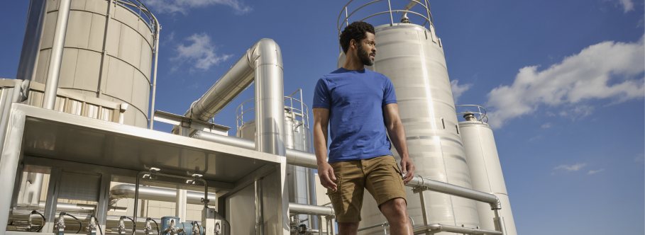 A man wearing a blue t-shirt and tan cargo shorts is standing at an industrial facility with large white storage tanks and piping under a blue sky.