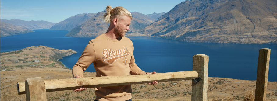 A man wearing a tan Strauss branded crewneck sweatshirt and tan cargo pants is sitting on a wooden fence at a scenic lakeside location with mountains and blue water in the background.