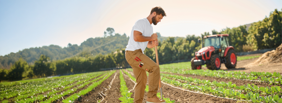 A man wearing a white t-shirt and tan double-front work pants is standing in a farm field with crop rows, holding a shovel, with a red tractor and trees in the background.