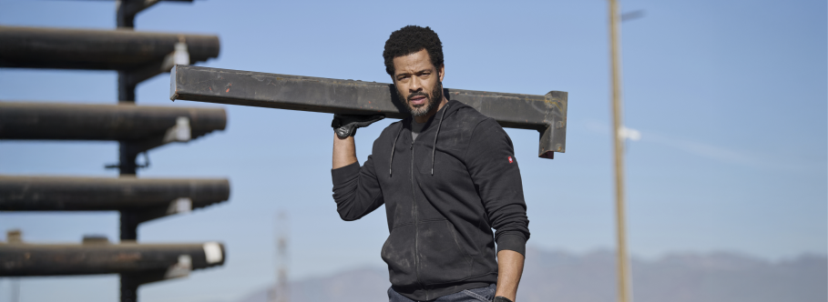 A man wearing a dark gray full-zip hoodie and jeans is carrying a metal beam on his shoulder at an outdoor construction site with wooden materials and blue sky.