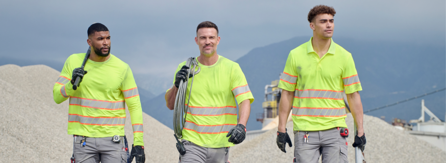 Three workers wearing neon yellow high-visibility shirts with reflective stripes, gray work pants, black gloves, and tool belts are walking at an industrial construction site with gravel piles and mountains in the background.