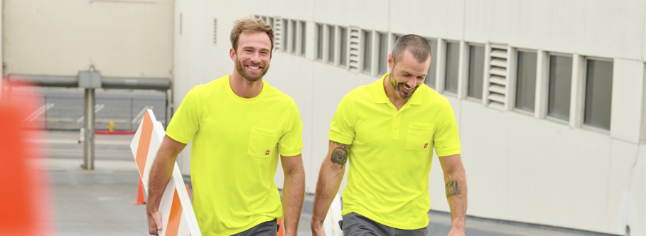 Two men wearing bright yellow high-visibility safety shirts walk together through an industrial warehouse area.