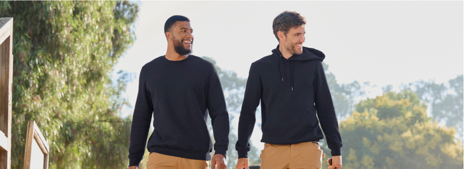 Two men wearing black sweatshirts and tan cargo pants are walking outdoors with trees and buildings in the background.