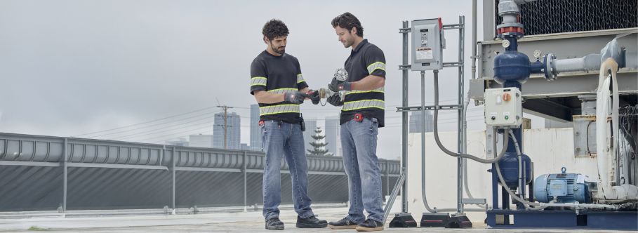 Two workers wearing blue work jeans and black shirts with high-visibility reflective stripes stand on a rooftop reviewing documents together.
