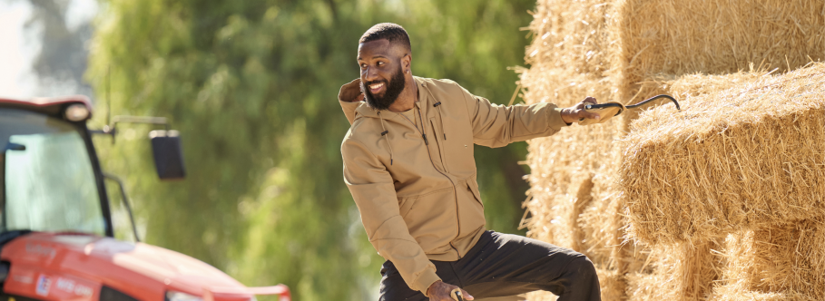A man wearing a tan lightweight work jacket and black cargo pants is sitting on hay bales at a farm with a red tractor and trees in the background.