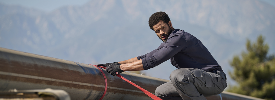 A man wearing a navy blue long-sleeve performance shirt and gray cargo pants  is securing a red strap on a metal structure at an outdoor work site.