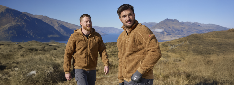 Two men wearing tan sherpa fleece jackets and cargo pants are walking outdoors in a scenic location with a lake, mountains, and grassland.