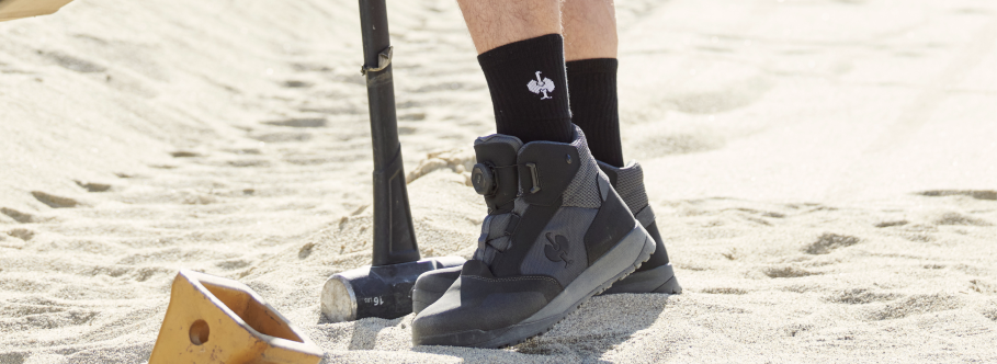 A person wearing black Strauss branded crew socks and graywork boots is standing on a sandy construction side.