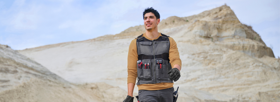 A man wearing a tool vest over a tan long-sleeve shirt stands outdoors in a sandy landscape with mountains behind him.