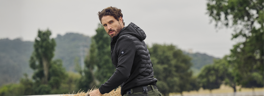 A man wearing a dark hooded work jacket stands in an outdoor field with trees and hills in the background.