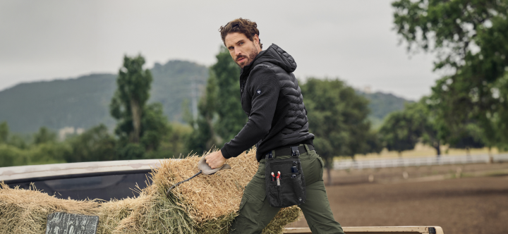 A man wearing a dark hooded work jacket stands in an outdoor field with trees and hills in the background.