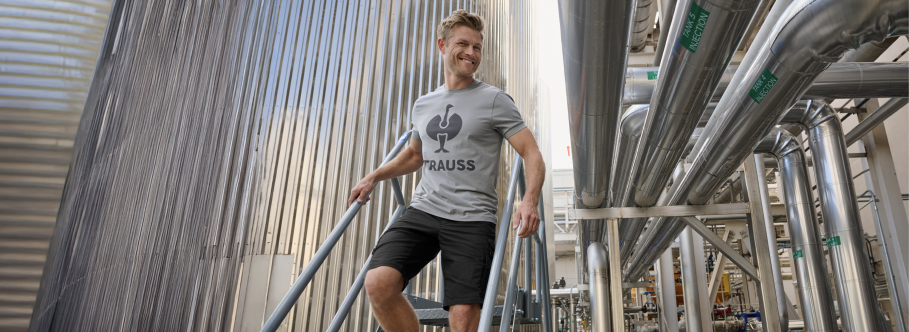 A man wearing a gray Strauss branded t-shirt and black work shorts is walking on metal stairs at an industrial facility with large metal silos and piping.