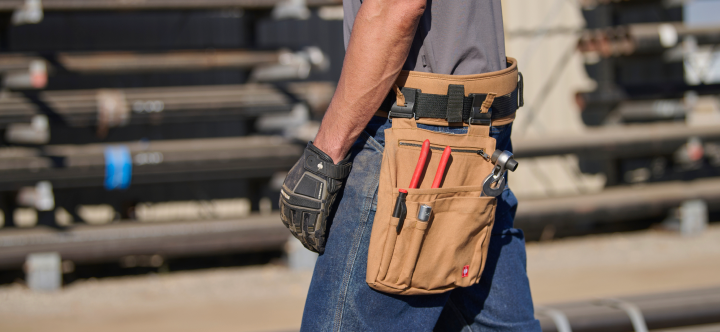 A worker wearing a tan canvas tool bag attached to a belt holds tools while standing at a construction site.