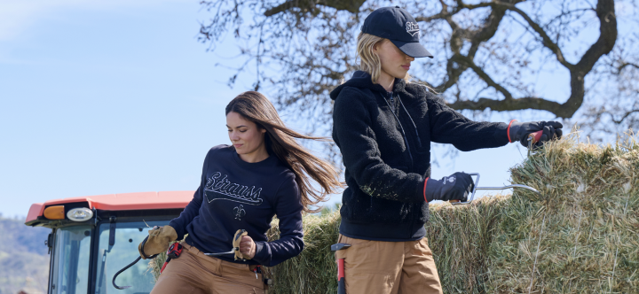 Two women wearing navy blue Strauss branded sweatshirts and tan cargo pants are working with hay at a farm with a tractor and trees.