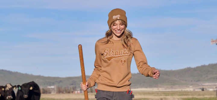 A woman wearing a tan branded crewneck sweatshirt and beanie walks through a field holding a walking stick.