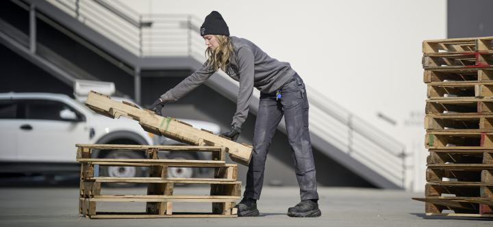 A woman wearing a black beanie, gray long-sleeve shirt, and dark work pants with reinforced double front fabric lifts wooden pallets at an industrial site.