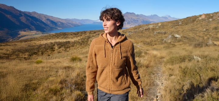 Person in a brown jacket standing on a trail with mountains and a lake in the background