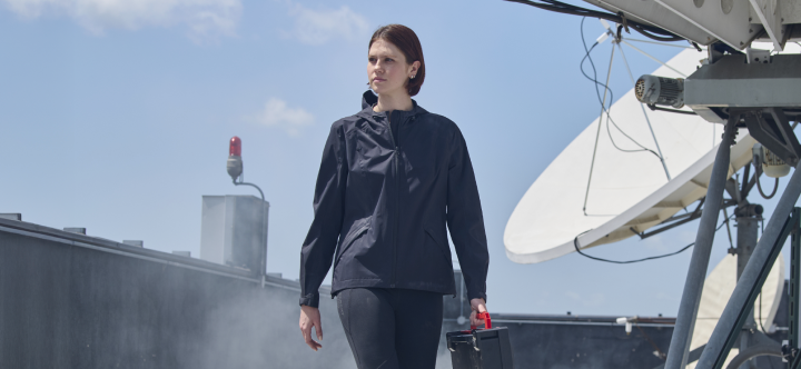 A woman wearing a dark lightweight windbreaker jacket walks on a rooftop with satellite equipment and cloudy skies in the background.