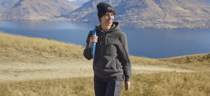 A woman wearing a dark fleece hoodie and beanie walks through grassy hills beside a mountain lake.