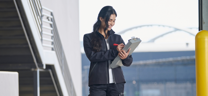 A woman wearing a dark softshell jacket with red accents reviews a tablet while standing on an airport walkway with aircraft visible in the background.