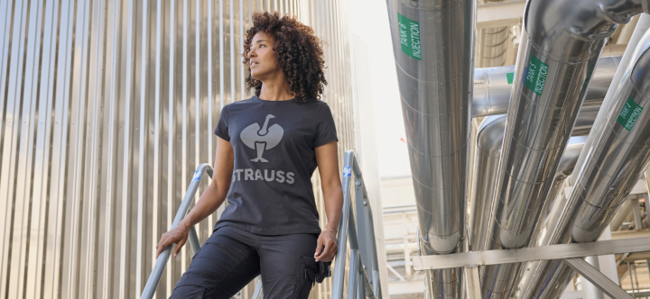 A woman wearing a branded work t-shirt and dark pants sits on industrial stairs in a modern factory setting