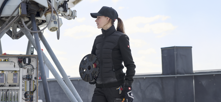 A woman wearing a black quilted work jacket with tool pockets and a black baseball cap stands on a rooftop with industrial equipment under a blue sky.