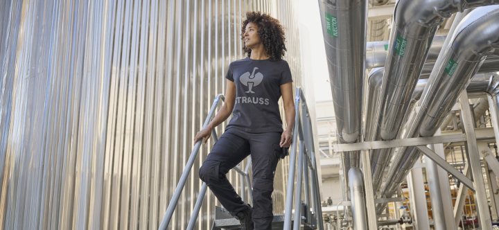 A woman wearing a branded work shirt and dark cargo pants walks down industrial metal stairs in a factory setting.