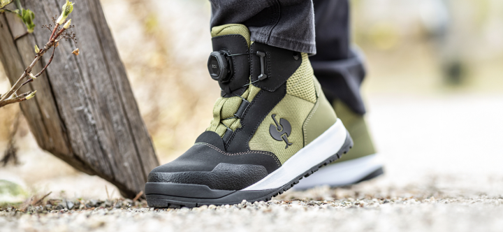 A close-up view shows a work boot with a BOA dial closure system in olive green and black worn outdoors on a gravel surface.