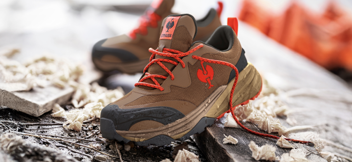 A pair of brown and black work shoes with red laces and Strauss branding sits on weathered wood with wood shavings, construction materials, and safety cones in the background.