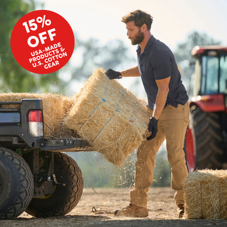A man in a navy polo shirt and tan work pants loads a hay bale onto the back of a pickup truck at a farm, with a red tractor in the background. A red circular '15% OFF' promotional badge is overlaid in the top left corner.