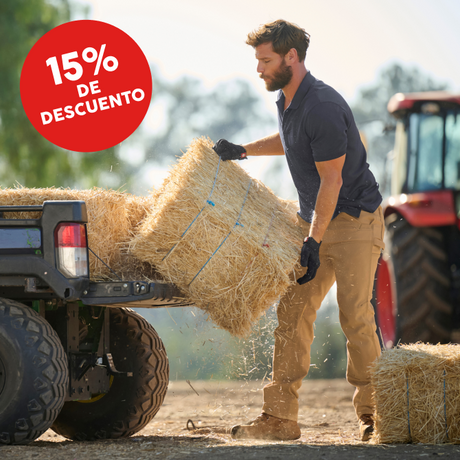 A man in a navy polo shirt and khaki pants loads a hay bale onto the back of a pickup truck at a farm, with a red tractor in the background. A red circular discount badge reading '15% DE DESCUENTO' is in the top-left corner.