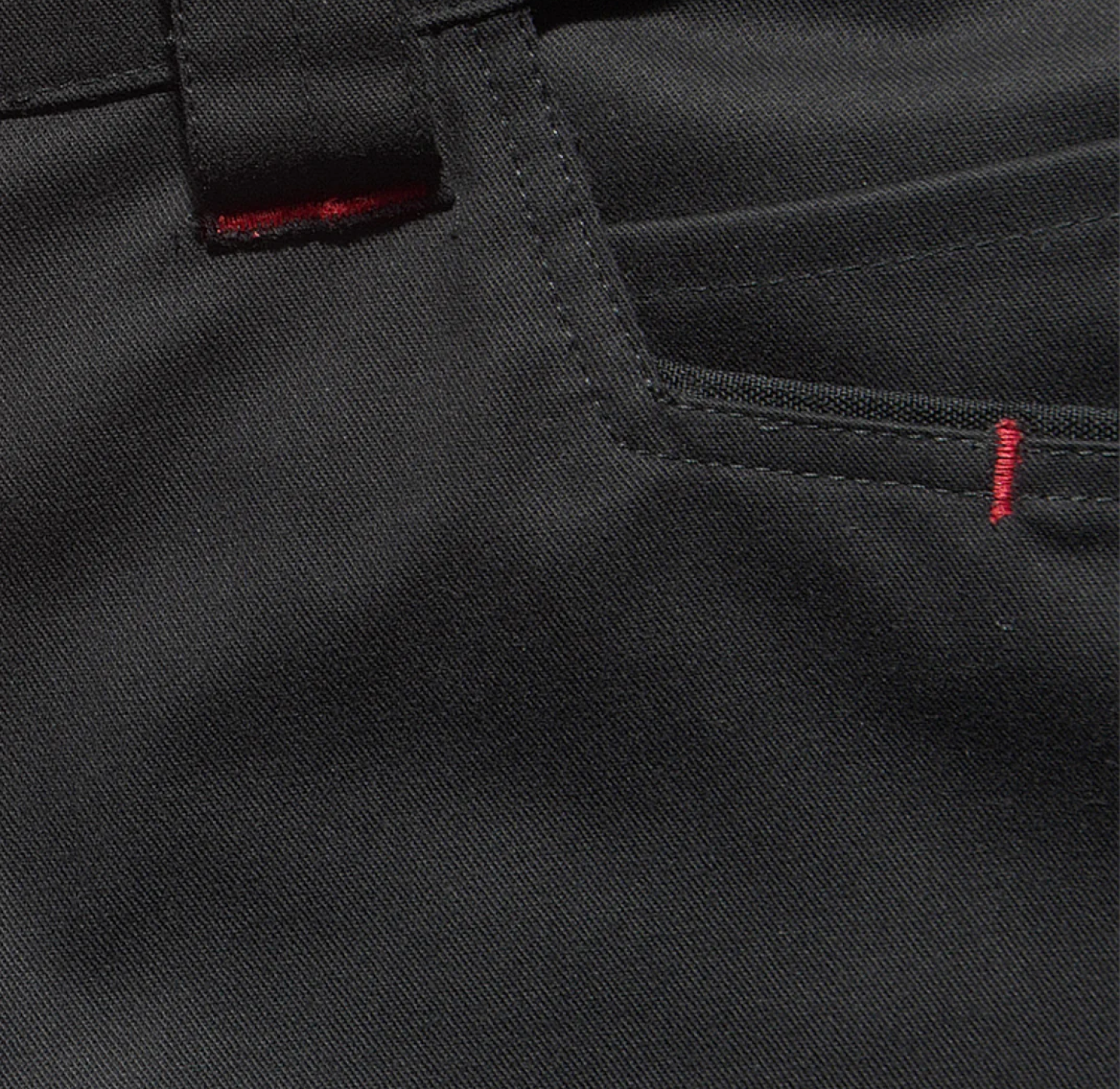 Close-up of black fabric pants with visible stitching and pocket. There are small red thread details on the belt loop and pocket corner, contrasting with the dark material.