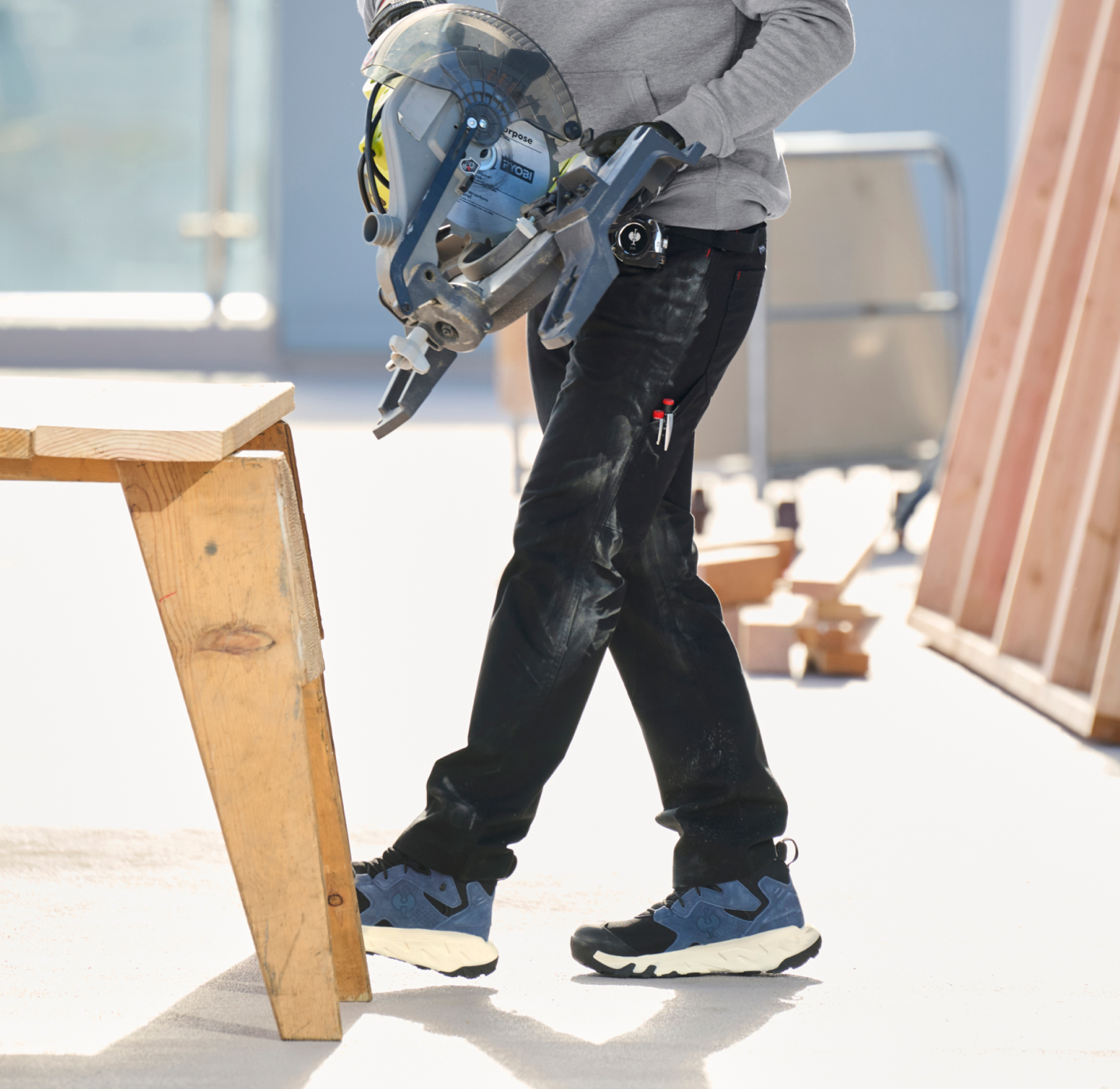 A person wearing blue sneakers and black pants holds a circular saw next to a wooden table in a bright indoor workspace with construction materials around.