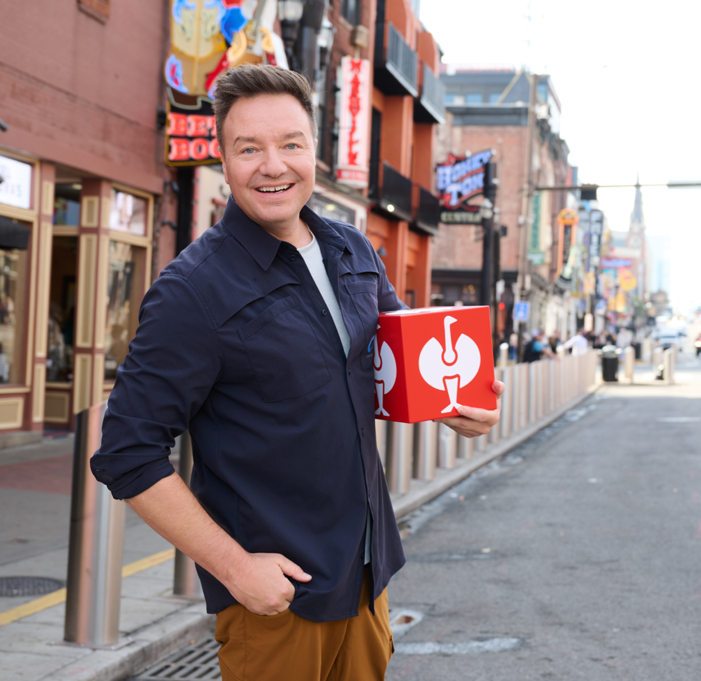 A smiling man stands on a city street holding a red and white box. Neon signs and storefronts line the busy sidewalk behind him.