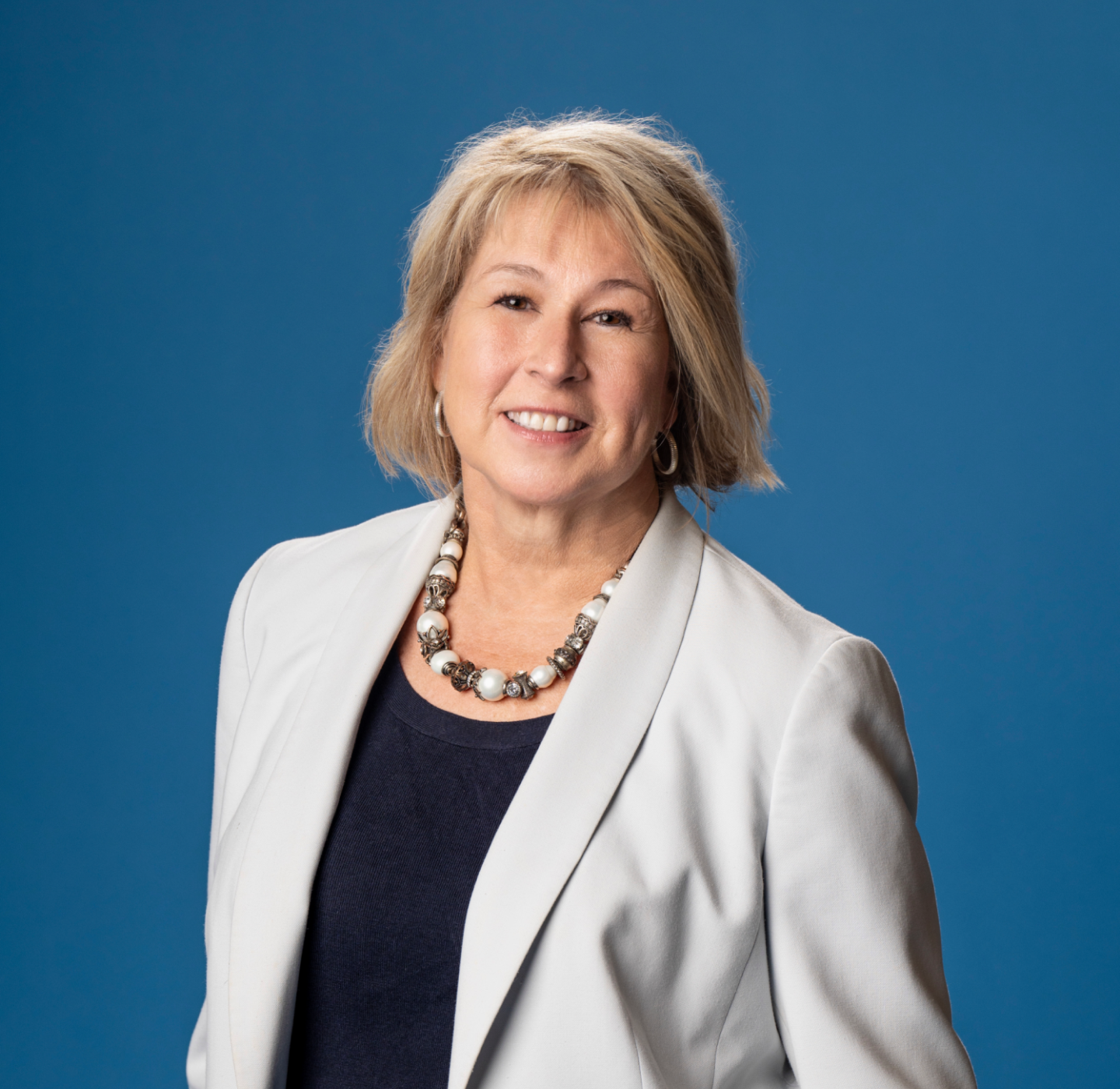 A woman with short blonde hair, wearing a light gray blazer, dark top, and a chunky necklace, smiles while standing in front of a solid blue background.