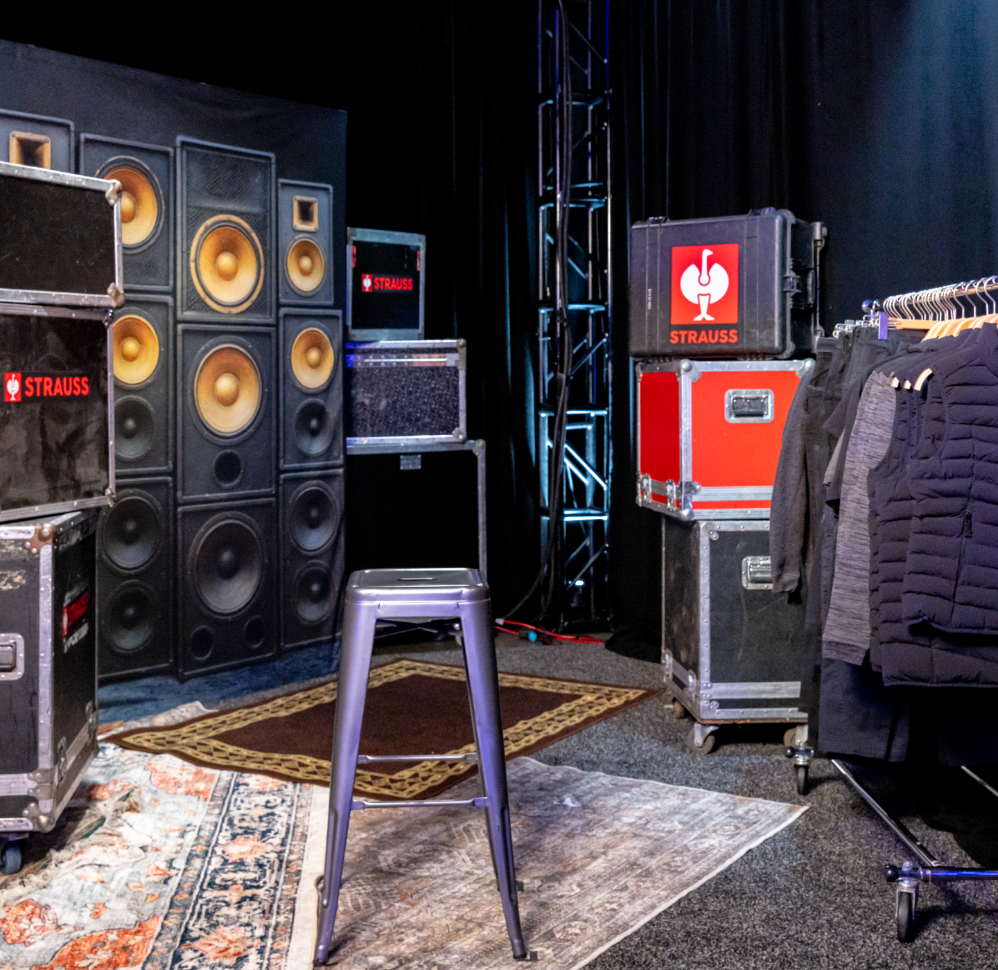 A metal stool sits on a rug in front of large speakers, surrounded by equipment cases and a rack of black jackets, in a backstage or studio setting with dark curtains.