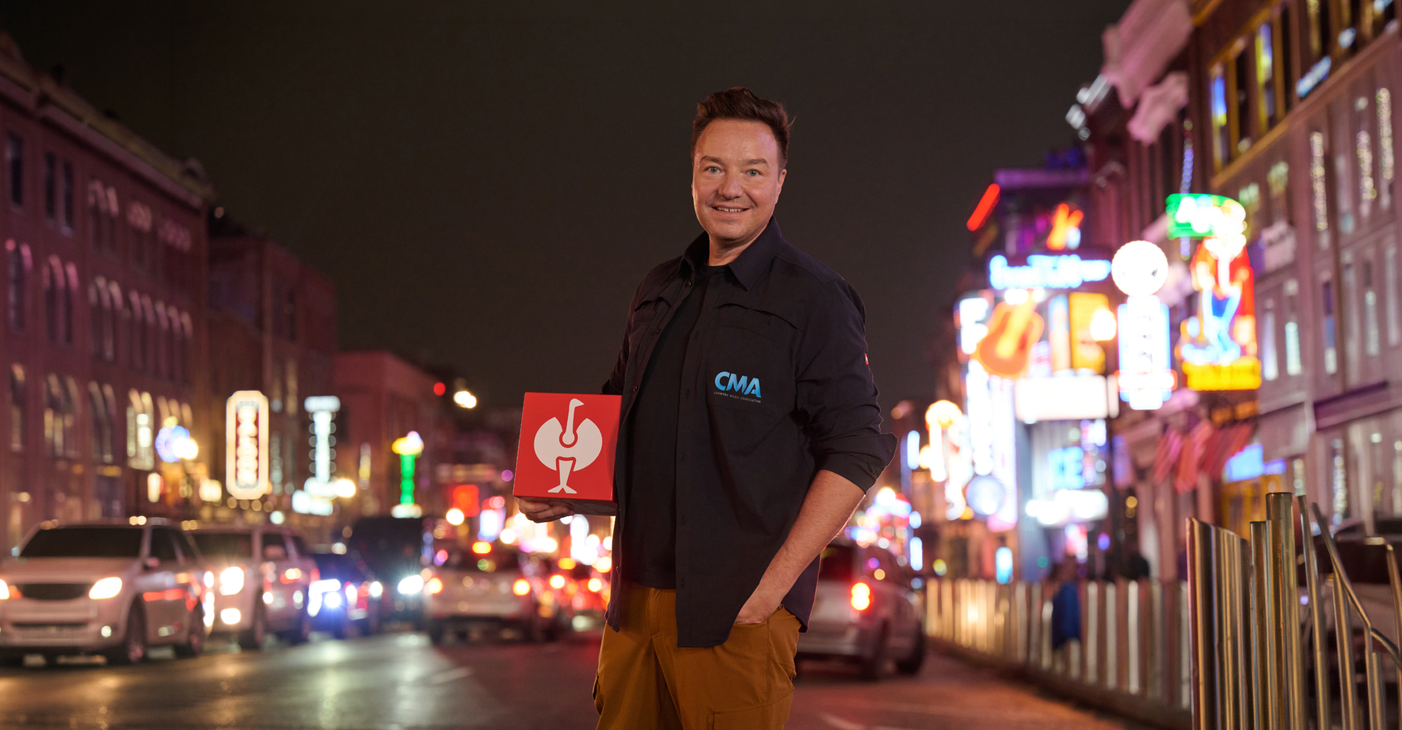 A man smiles at the camera while standing on a busy city street at night, holding a red and white box. Colorful neon signs and car headlights light up the background.