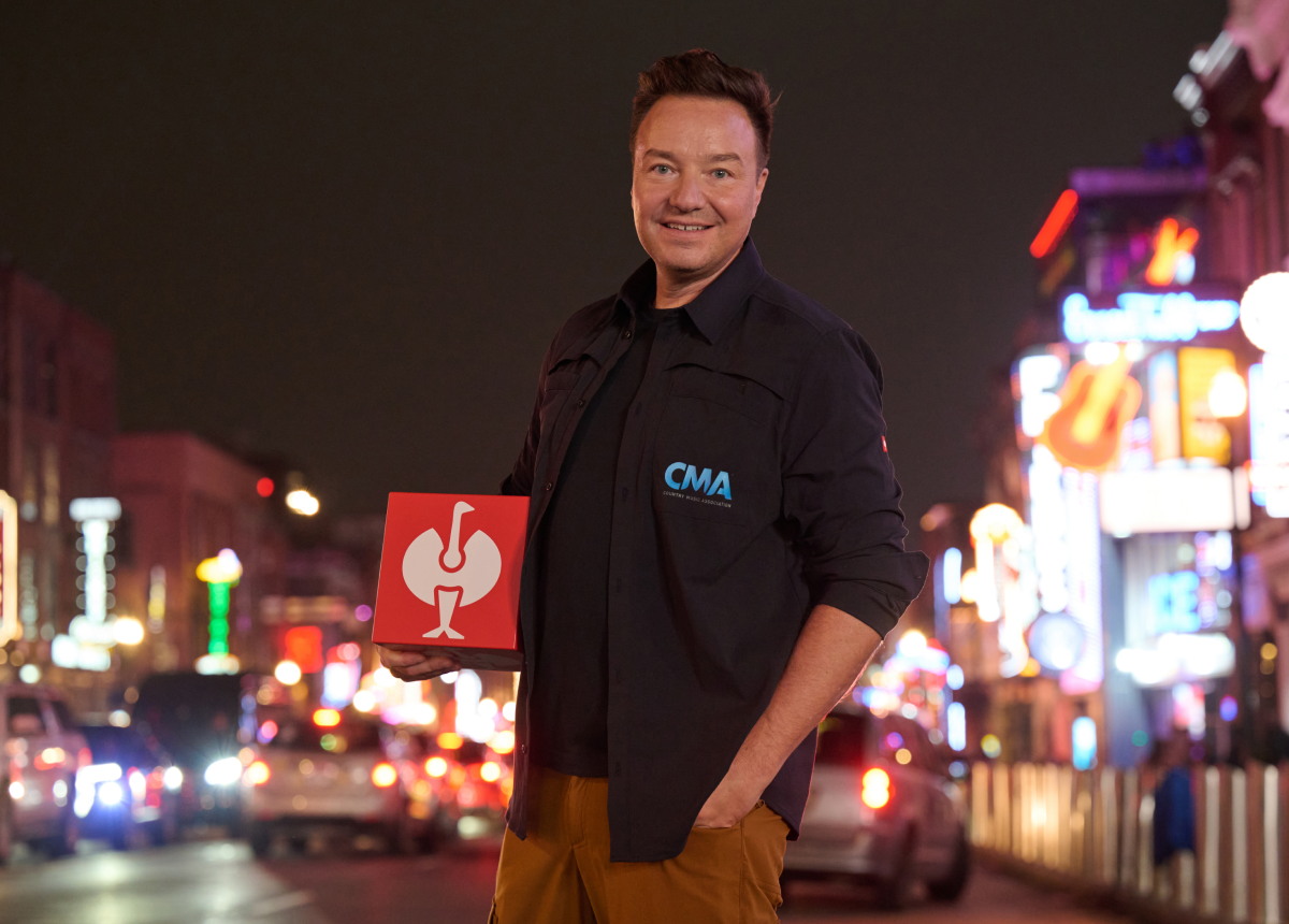A man stands on a brightly lit city street at night, smiling and holding a red box with a white logo. He wears a dark jacket with CMA on the chest. Neon signs and blurred car lights fill the background.