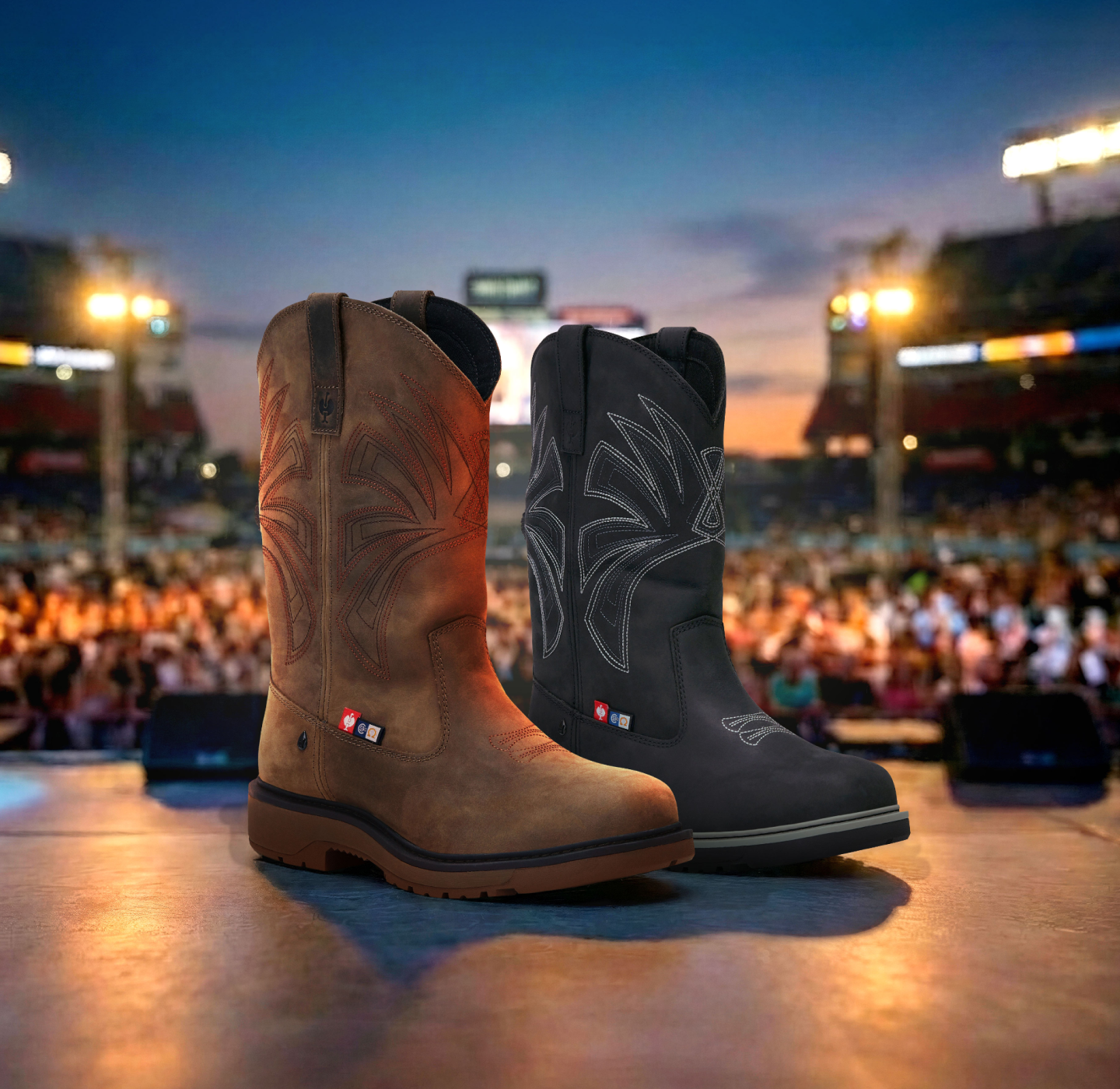 Two pairs of cowboy boots, one brown and one black, are displayed on a stage with bright lights. In the blurred background, a large crowd is gathered at an outdoor event or concert.