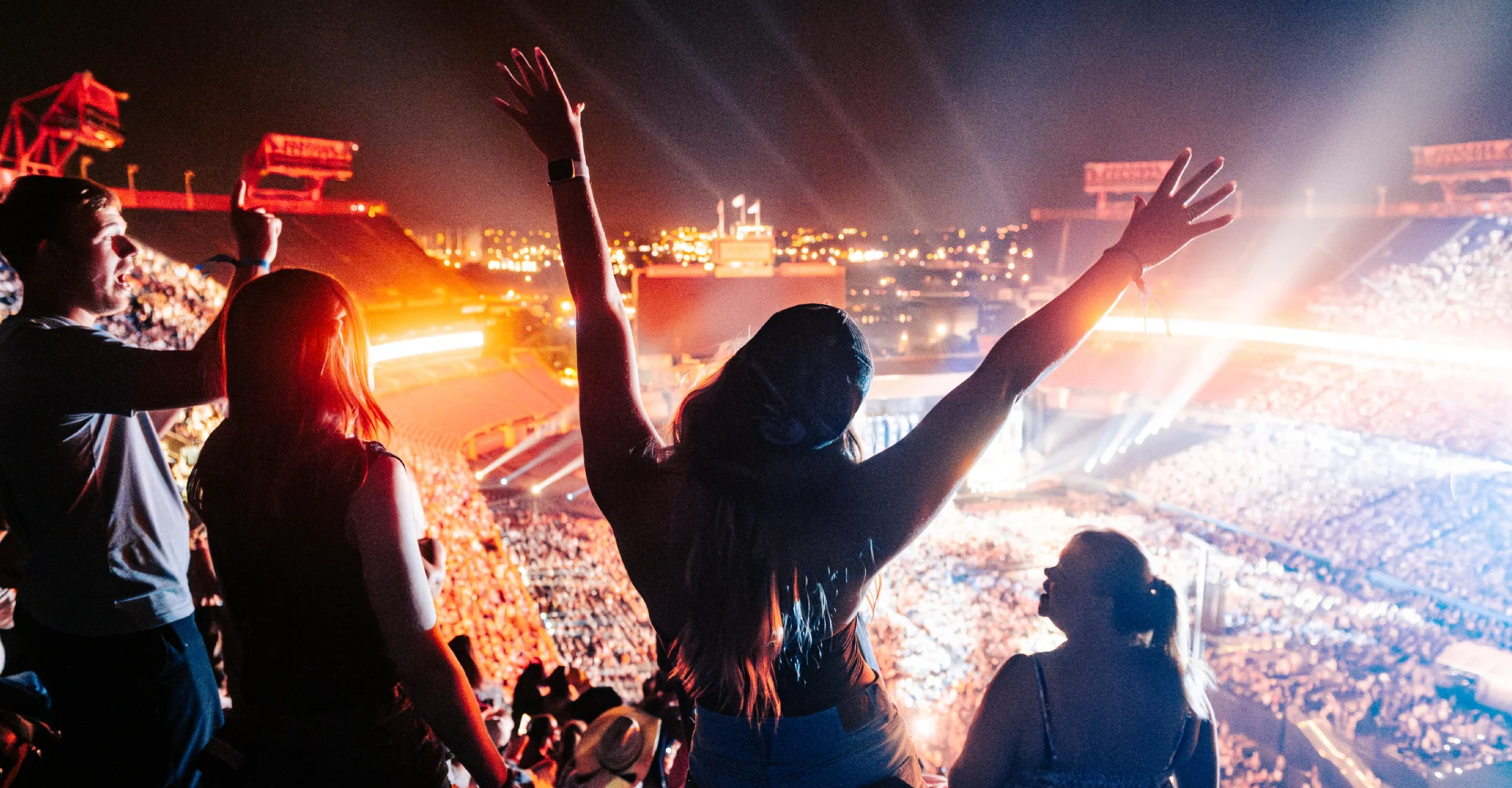 A crowd enjoys a nighttime stadium concert. In the foreground, a person stands with arms raised, surrounded by others. Bright lights illuminate the stage and thousands of fans fill the seats.
