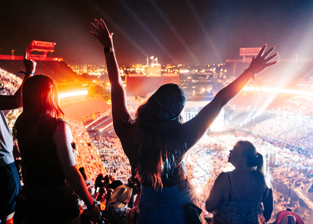 A crowd at a stadium concert at night, with a person in the foreground raising their arms in excitement as lights and people fill the venue.
