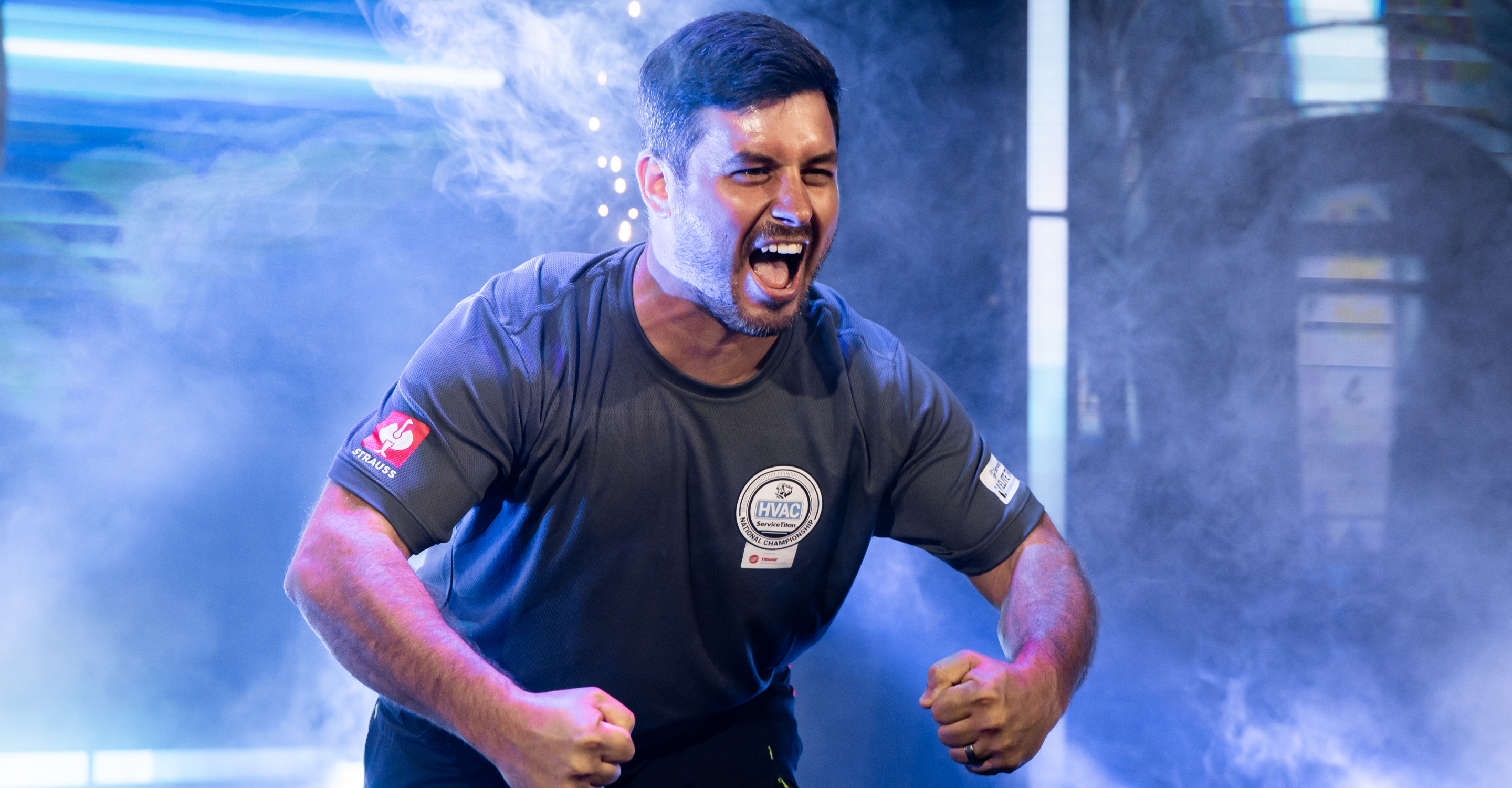 A man in a sports shirt clenches his fists and yells passionately, surrounded by dramatic smoke and colorful lighting on stage.