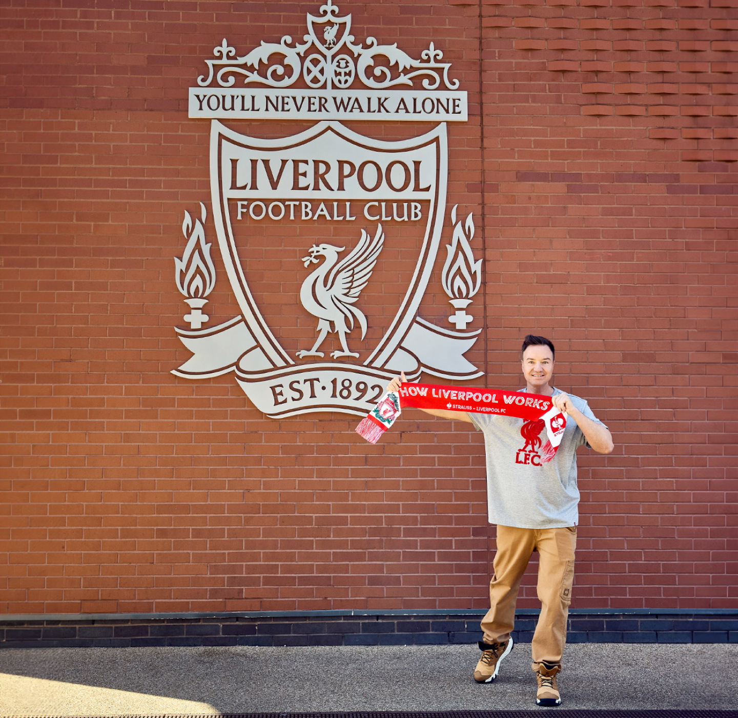 A person stands in front of a large Liverpool Football Club crest on a brick wall, holding a red Liverpool scarf and smiling at the camera.