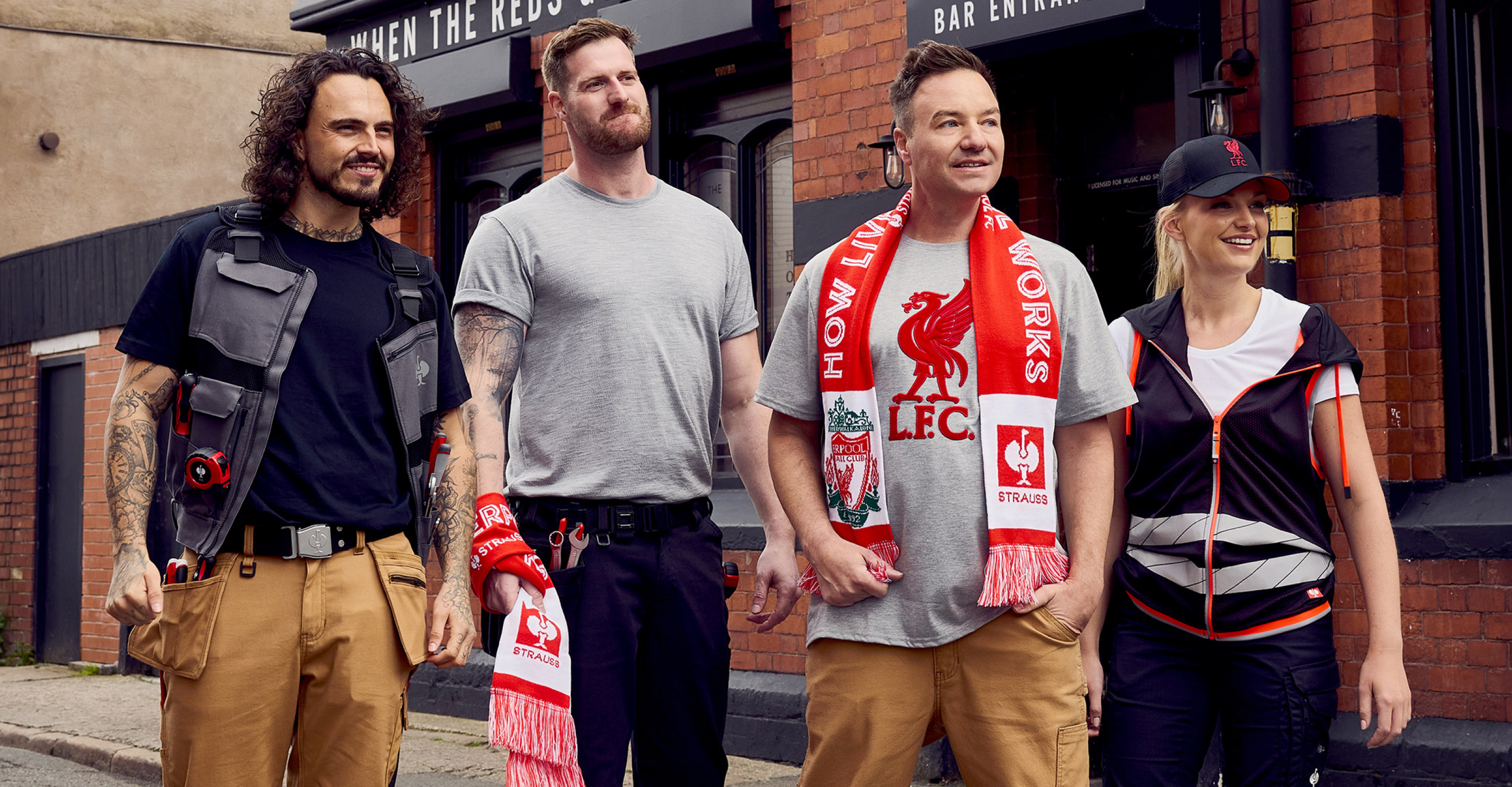 Four people stand outside a brick building, two wearing Liverpool FC scarves, smiling. They are dressed casually, with two in work vests and two in T-shirts. The mood appears friendly and relaxed.