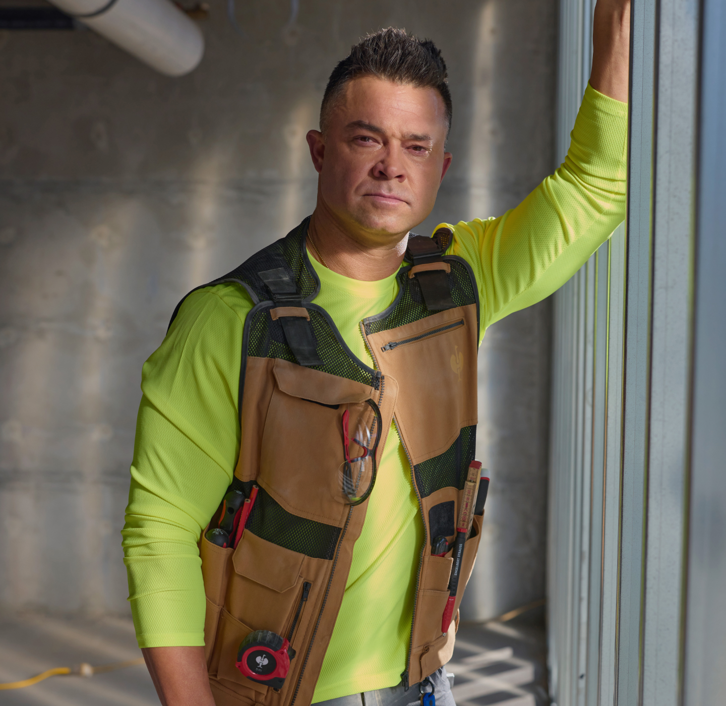 A man in a yellow shirt and tool vest stands by a window in a construction setting, looking at the camera. Tools are visible in his vest pockets, and sunlight streams through the glass.