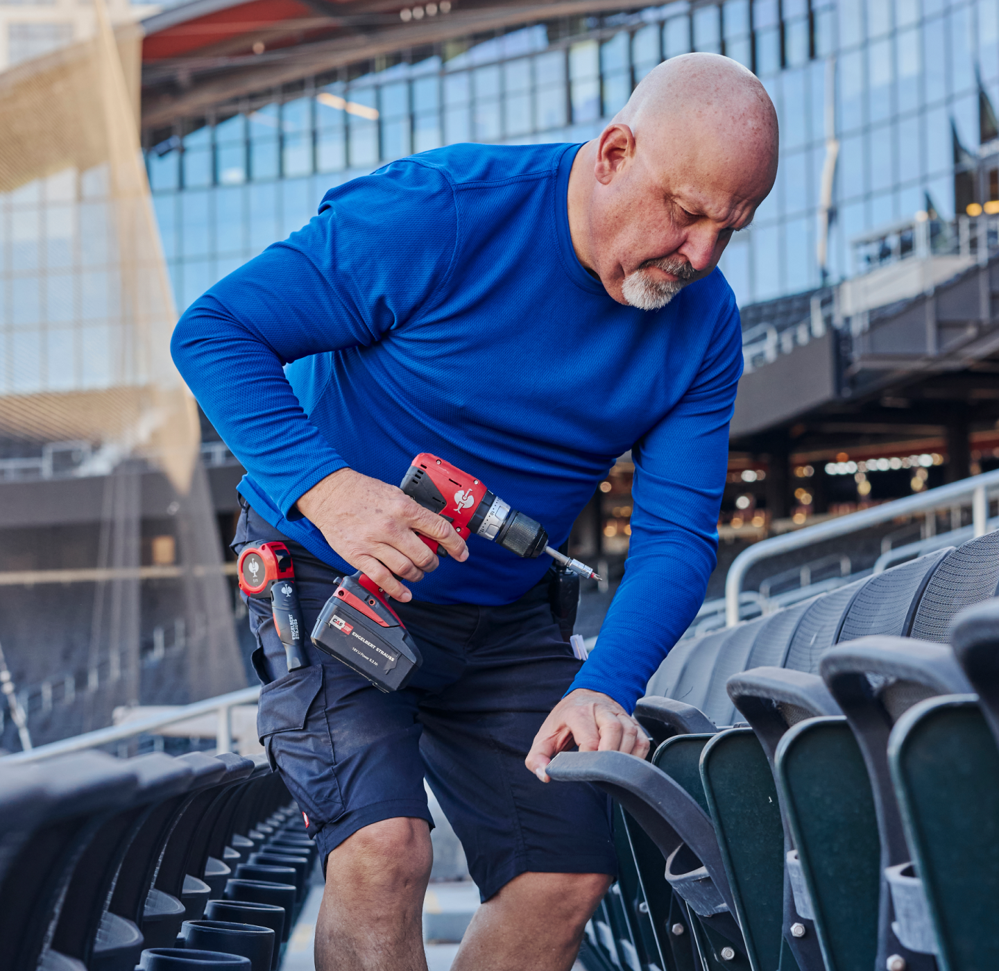 A man in a blue shirt uses a cordless power drill to fix or assemble stadium seats. He is focused on his task, surrounded by empty rows of seats and modern glass buildings in the background.